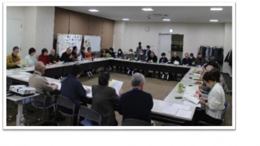 ロの字に設置された長机に関係者が座り、沢山の人達が資料に目を通している室内を写した写真