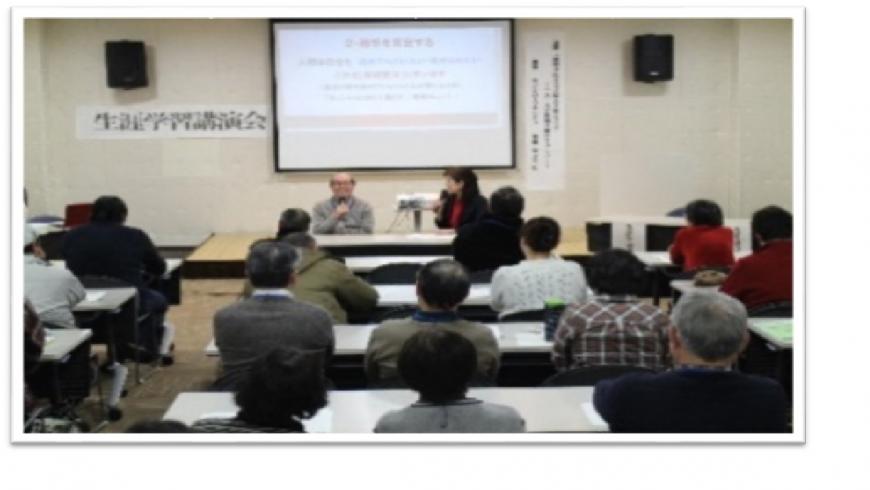 スクリーンの前に座っている男性がマイクを持ち、参加者の人たちに説明をしている写真