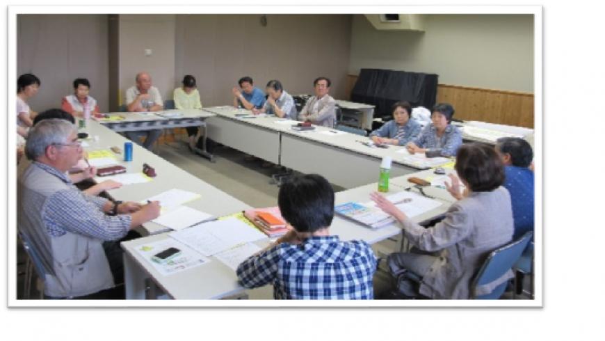 長机をロの字に設置した室内で中央の女性が身振り手振りで話をしている様子を参加者の人達が聞いている写真