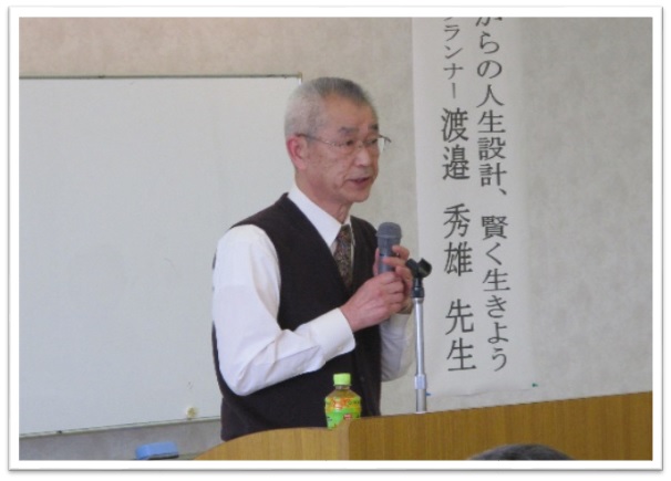 檀上に立つ黒色のベストを着た男性がマイクを持って話をしている写真
