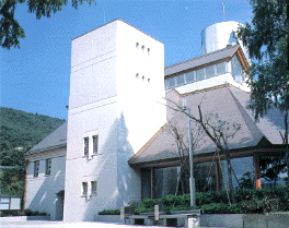 白い長方形の建物と焦げ茶色いの屋根の後ろに銀色の筒状の塔がある古関裕而記念館の外観写真