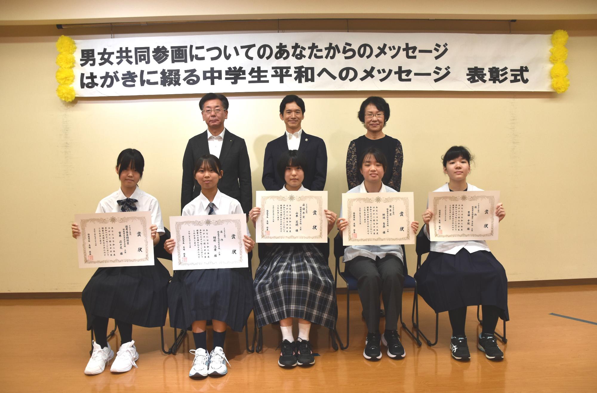 前列にイラスト部門入賞者の生徒5名が賞状を持って椅子に座り、後方に市長と関係者の方2名が並び写っている記念写真