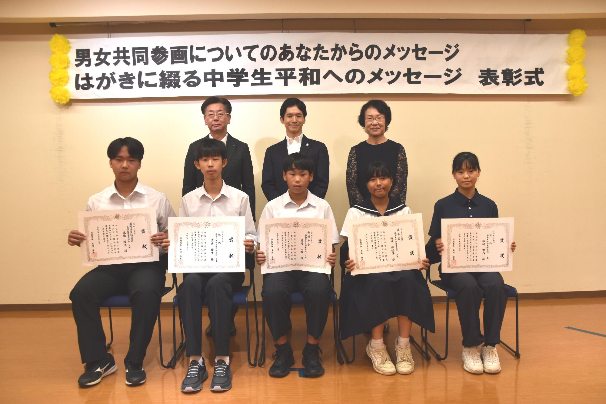 前列にメッセージ部門入賞者の生徒5名が賞状を持って椅子に座り、後方に市長と関係者の方2名が並び写っている記念写真
