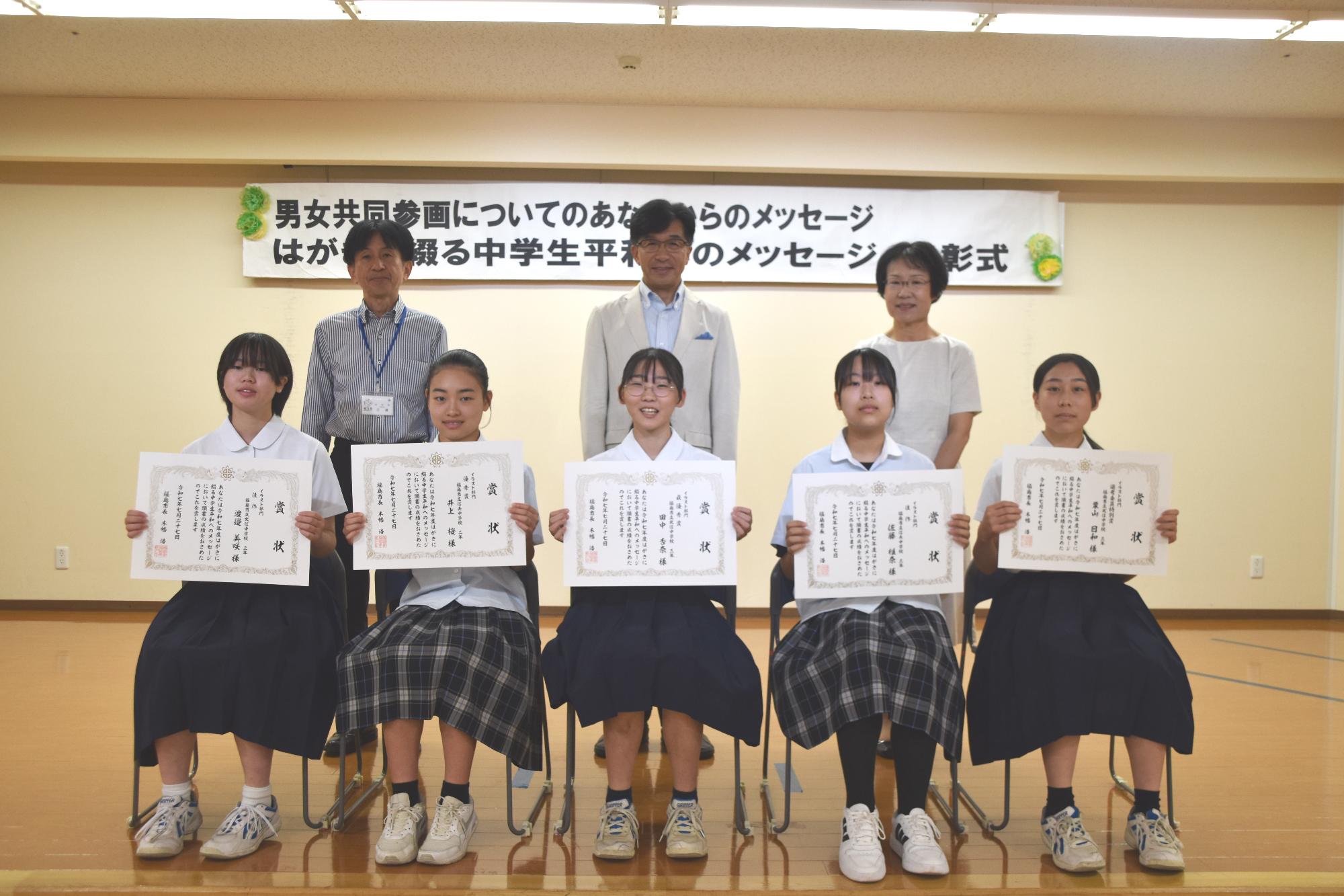 前列にイラスト部門入賞者の生徒5名が賞状を持って椅子に座り、後方に市長と関係者の方2名が並び写っている記念写真