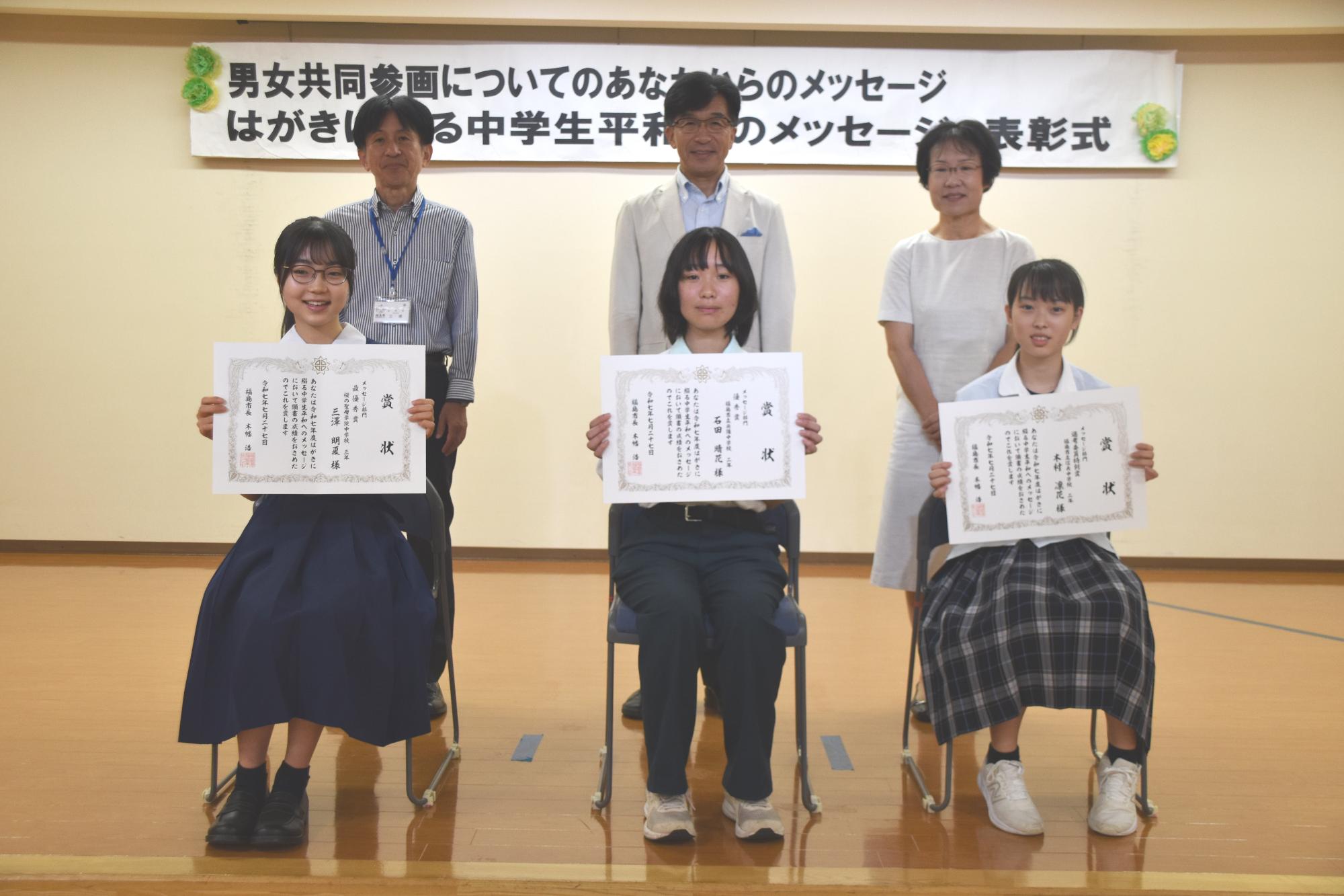 前列にメッセージ部門入賞者の生徒3名が賞状を持って椅子に座り、後方に市長と関係者の方2名が並び写っている記念写真