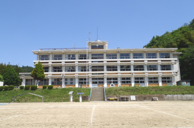 グラウンドと法面を挟んで一段高くなった敷地に建つ、3階建ての校舎の施工前全景写真