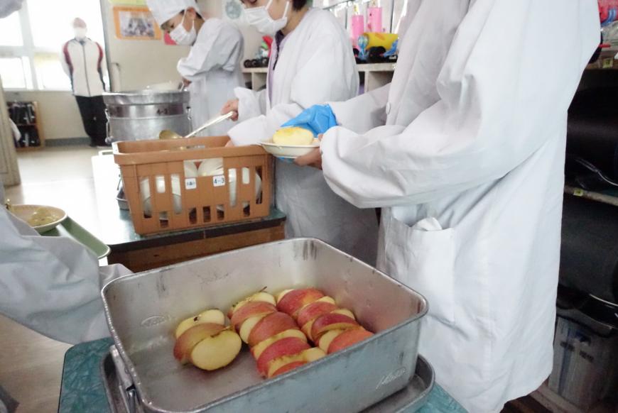 シルバーの色をしたバットにカットされたリンゴがあり、給食当番の人がお皿に配膳している写真