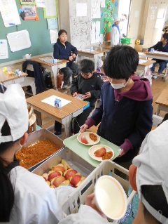 教室内で、給食着を着た生徒からカットされたリンゴを受け取る生徒の写真