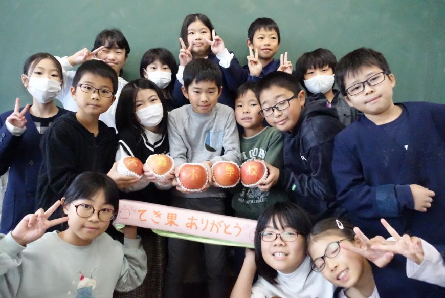 黒板の前に集まった生徒達がリンゴを囲んで記念撮影をしている写真