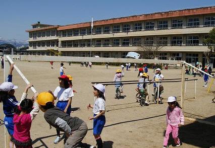 オレンジ色の屋根の3階建ての渡利小学校校舎の前の校庭で、たくさんの生徒たちが、倒したサッカーゴールや一輪車など思い思いに遊んでいる様子の写真