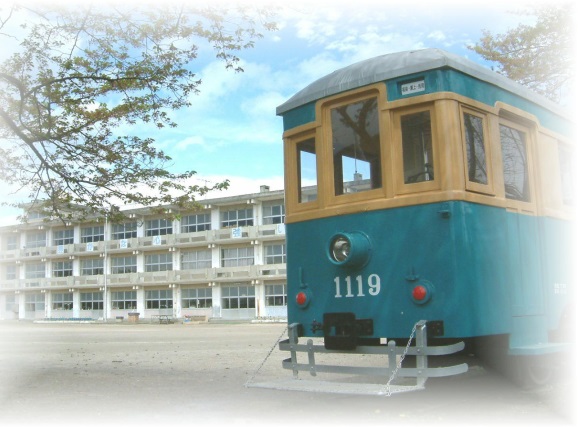3階建ての校舎の前にある校庭の脇に、黄色と水色の配色の車体の正面の「1119」と番号がペイントされた電車が置かれている瀬上小学校の外観写真