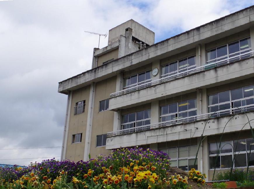 紫や黄色の花が咲き誇った花壇越しに3階建ての校舎を写した、月輪小学校の外観写真