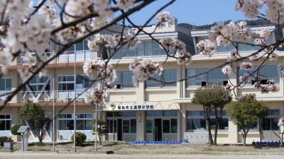 花が咲いた桜の枝越しに写した3階建ての湯野小学校の外観写真