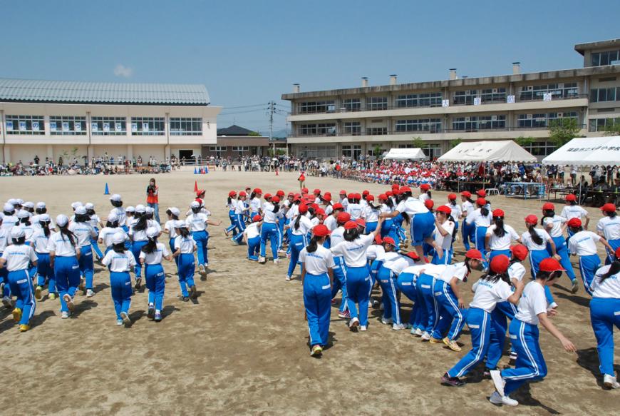 大森小学校の3階建ての校舎と体育館の手前の運動場で体操着を着て赤と白の帽子をかぶったたくさんの生徒たちが集まっている写真