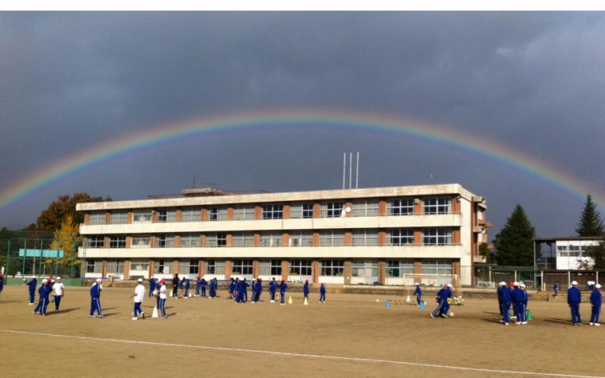 空に出ている虹のアーチの真ん中に北沢又小学校の3階建ての校舎があり、手前の運動場で体操着を着た生徒たちが活動している写真