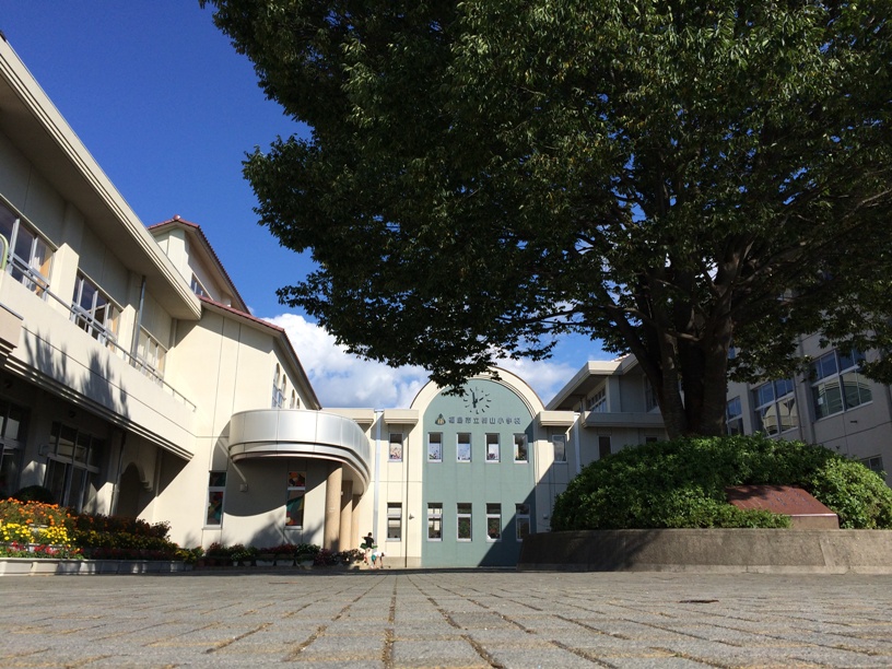 大きな木が生えた植栽の左に2階建ての校舎、奥に時計のついた緑色の壁の建物がある御山小学校の写真