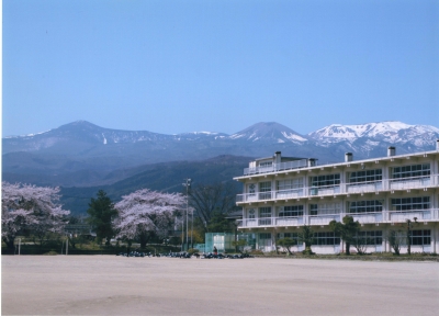 雪の残る山並みを背景に建つ西信中学校の3階建ての校舎をグラウンドから撮った写真