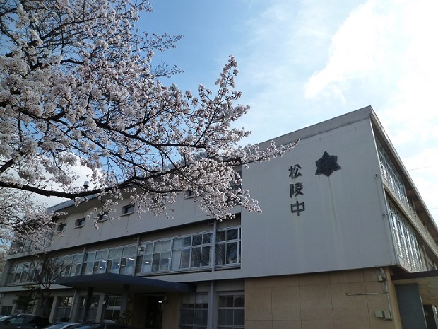 花をつけた桜の枝越しに撮影した、白い壁に「松陵中」の文字がついた松陵中学校の校舎の写真