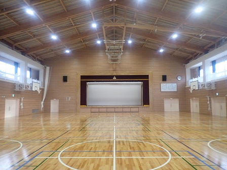 奥の壁に舞台が設けられ、左右の壁にバスケットゴールが設置された、板張りの余目小学校屋内運動場の内観写真