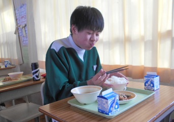 机の上に乗った給食をおいしそうにほおばっている男子生徒の写真