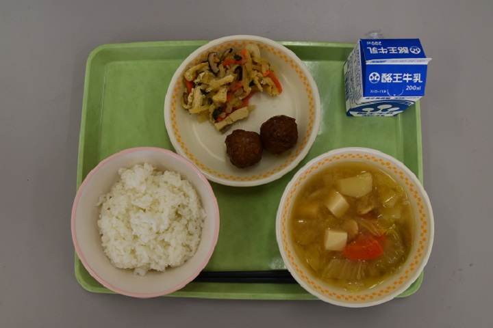 若草色のトレーの上に、ごはん、打ち豆のみそ汁、きんぴら肉団子とひきな炒りが盛り付けられた皿、パックの牛乳がのっている写真