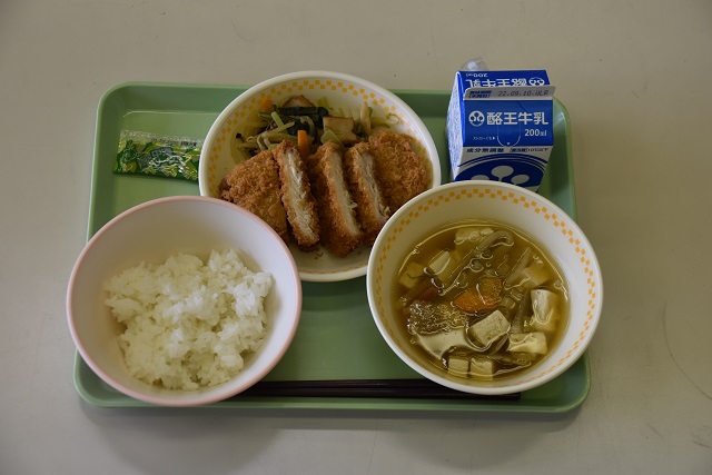 若草色のトレーの上に、ごはん、けんちんみそ汁、トンカツと小松菜の炒め煮が盛り付けられた皿、パックの牛乳が乗っている写真