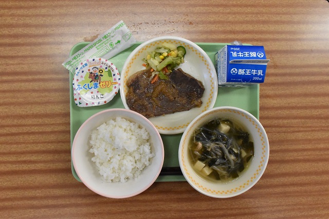 若草色のトレーの上に、ごはん、五目スープ、県産牛ステーキおろしソースかけとブロッコリーサラダが盛り付けられた皿、パックの牛乳、県産りんごゼリーが乗っている写真