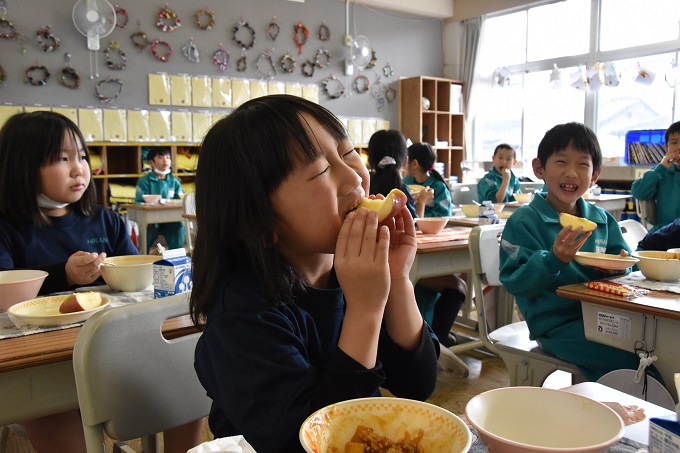 教室でみんなが給食を食べる中、おいしそうにリンゴにかじりつく女子児童の写真