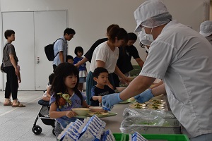 手前で女の子が給食白衣と帽子、マスクを身に着けた男性から、手に持った給食が乗ったトレーにパック牛乳を乗せてもらっており、その横では参加者の親子が順番に給食を受け取っている様子の写真