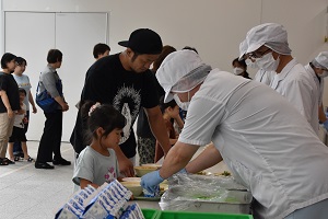 手前で給食白衣と帽子、マスクを身に着けた男性が女の子のトレーにパック牛乳を乗せ、その横では参加者の親子が順番に給食を受け取っている様子の写真