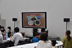 「給食ができるまで」の資料が映し出された大きなモニターを見ている参加者たちの写真