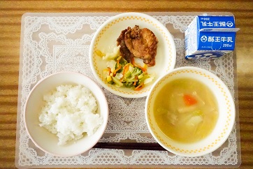 白いレース模様のランチョンマットの上に、ごはん、野菜のかす汁、鶏のから揚げレモンソースかけとブロッコリーサラダが盛り付けられた皿、パックの牛乳が乗っている写真