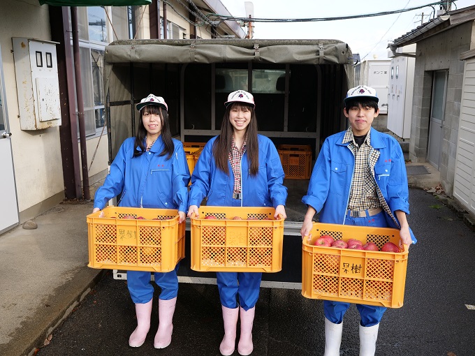 青い作業着を着た3人の高校生がリンゴがたくさん入った黄色のコンテナを持ち、横一列に立っている写真
