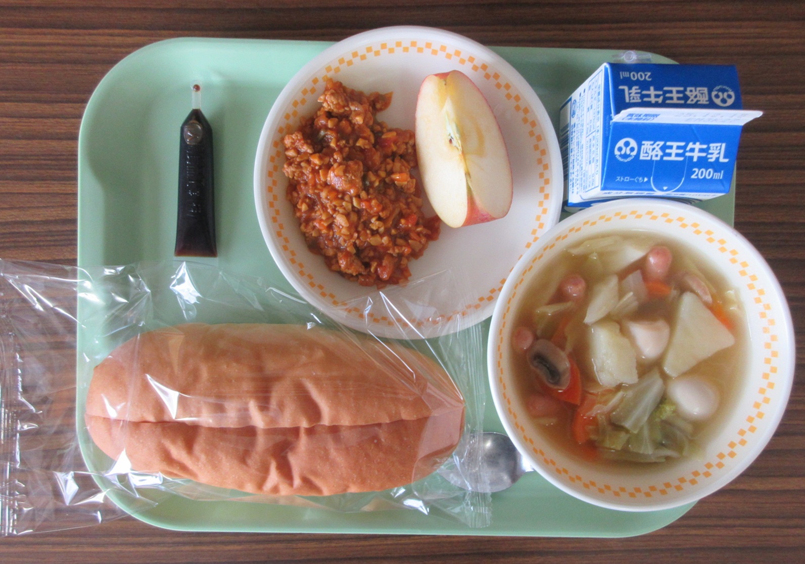 お盆に切り目入りコッペパン、豆のドライカレー、ポトフ、りんご、牛乳、ミルメークが配膳された給食の写真