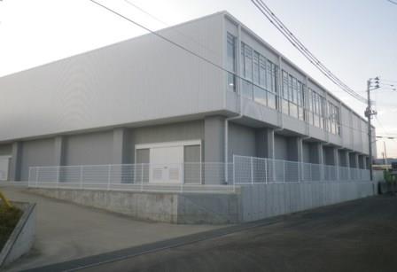 コンクリートの壁と白いフェンスで道路と隔てられたグレーの外壁の四角い西信中学校屋内運動場の外観写真