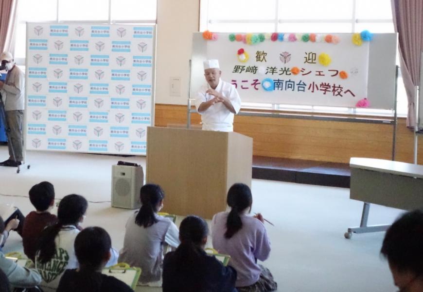 「歓迎 野崎洋光シェフ ようこそ南向台小学校へ」と書かれた紙が貼られたホワイトボードの前の演台に立ち、床に座っている生徒達に話をしている野崎洋光氏の写真