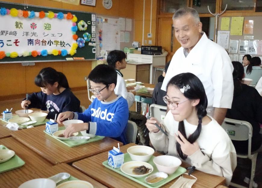 席に座り給食を食べている生徒達の様子を見ている野崎洋光氏の写真