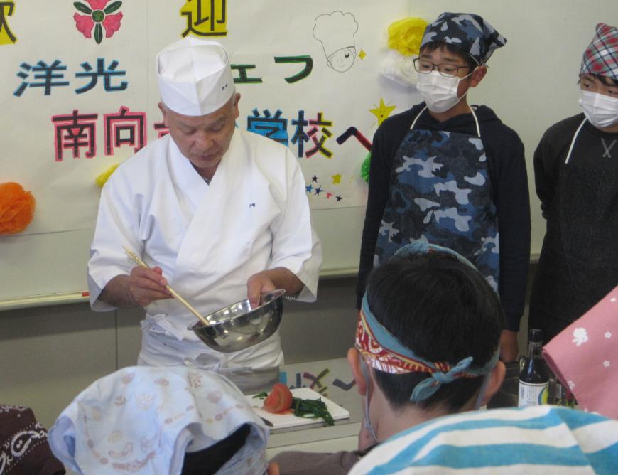 白い調理着を着た野崎氏が菜箸とボールを持ち調理をしている様子を生徒達が真剣に見ている写真