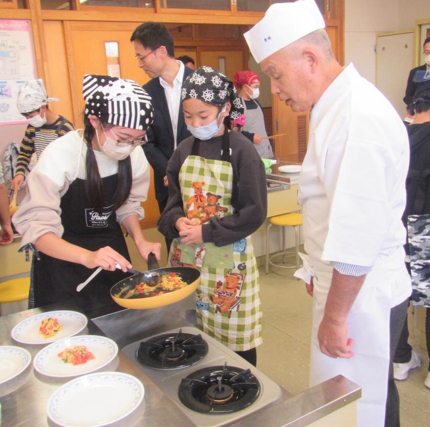 1人の女子生徒がフライパンで食材を炒めている様子を見守っている野崎氏の写真