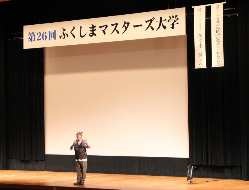 第26回ふくしまマスターズ大学の横断幕が飾られた舞台の中央にたって講演をしている佐々木洋さんの写真