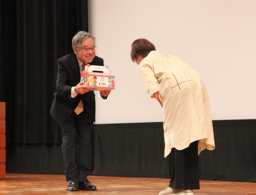 三宅民夫さんと女性が挨拶をしながら贈呈品を受け取っている記念品贈呈の写真