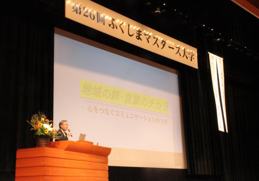 第26回ふくしまマスターズ大学の横断幕が飾られた演台で三宅民夫さんがスクリーンを使いながら講演をしている写真