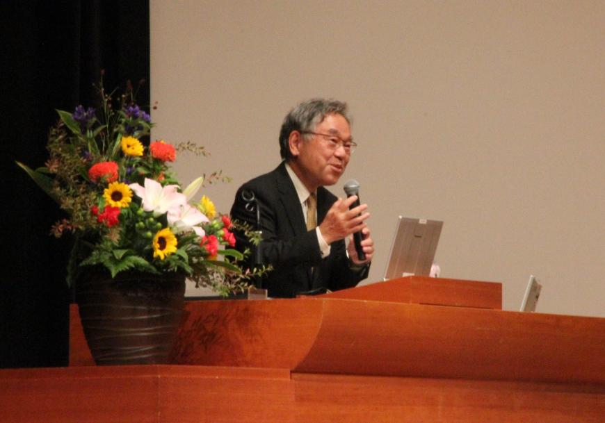 黄色や白、オレンジなどの花が花瓶に飾られた演台でマイクを持っている三宅民夫さんをアップで写した写真