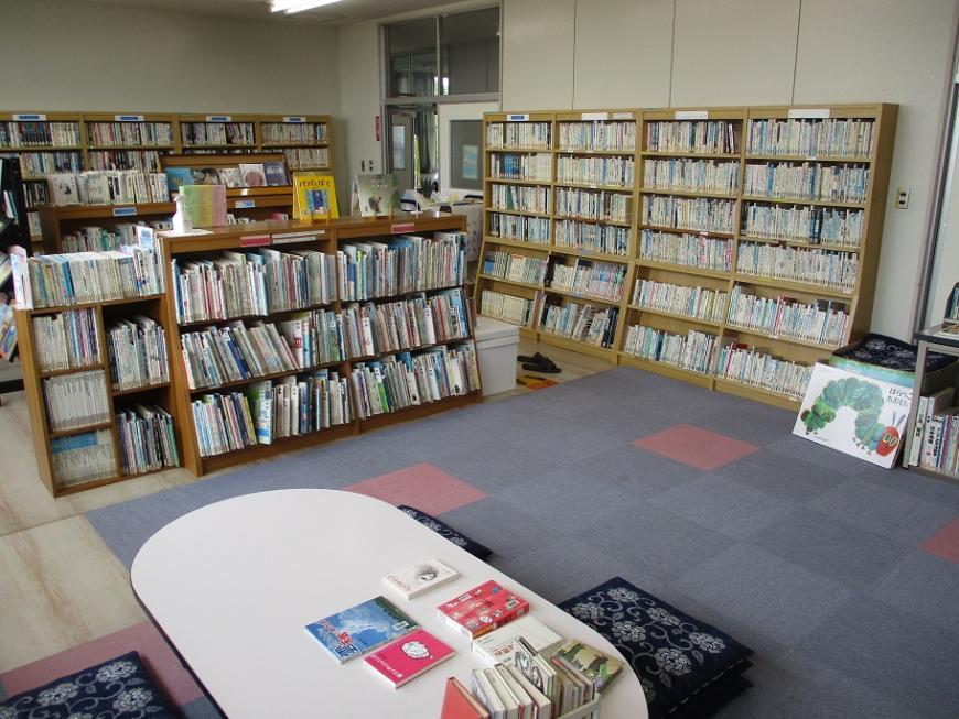 手前には楕円形の机が置かれ、奥の壁、中央の仕切りの箇所の本棚にたくさんの本がおかれている図書室の写真