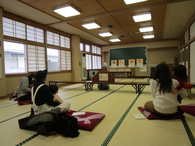 畳に座布団がひかれ子供をひざにのせたお母さんが座り前方の紙芝居をみている写真