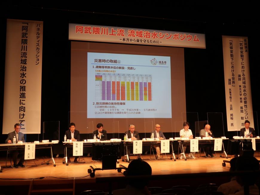 「阿武隈川上流流域治水シンポジウム」と書かれた横断幕が貼られ、スクリーンに映像が映し出された舞台の上に長机といすが設置され、市長や関係者が座っている