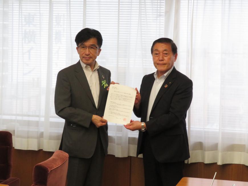 白いレースカーテンがかかった窓際で市長と男性が一緒に用紙をもち記念撮影している写真