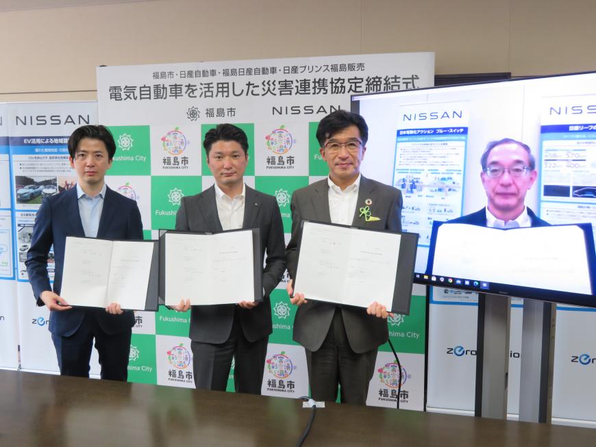 福島市のバックボードの前に置かれた男性が写ったスクリーンの横に、市長と男性2名が協定書を手に持って立ち記念撮影している写真
