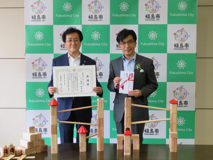 福島市のバックボードの前で目録の封筒を持った市長と感謝状を持った男性が並び、その前に設置された長机に木製の知育玩具が置かれている写真