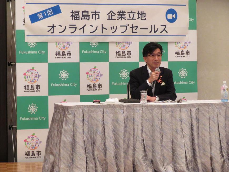 「第1回福島市企業立地オンライントップセールス」と書かれた横断幕が貼られた福島市のバックボードの前に設置された席に座った市長がマイクで話をしている写真